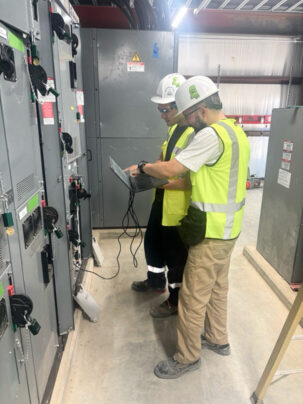 Two electrical engineers in safety gear using a laptop to perform system diagnostics on industrial control panels.